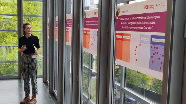Eine Person steht in einem modernen Raum vor mehreren Informationsplakaten. Die Plakate thematisieren Barrierefreiheit im öffentlichen Raum und deren Einfluss auf Mobilität und Chancengleichheit. Es gibt Grafiken und Texte, die die unterschiedlichen Zugänglichkeiten veranschaulichen.