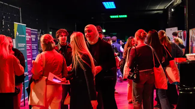 Eine Gruppe von Menschen steht in einem Raum und kommuniziert lebhaft. Einige halten Informationsmaterial in den Händen. Im Hintergrund sind Plakate und Stände sichtbar, die auf eine Veranstaltung hinweisen. Die Atmosphäre ist gesellig und engagiert.