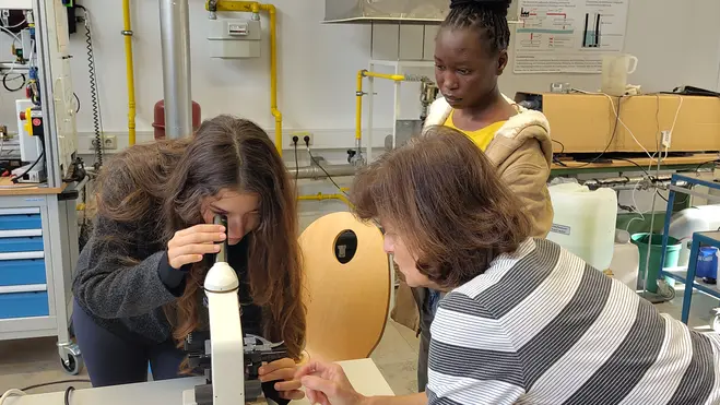 Drei Personen befinden sich in einem Labor. Eine junge Frau blickt durch ein Mikroskop, während eine andere Person neben ihr steht und zusieht. Eine dritte Person hilft, indem sie auf das Mikroskop zeigt. Die Atmosphäre ist lehrreich und experimentell.