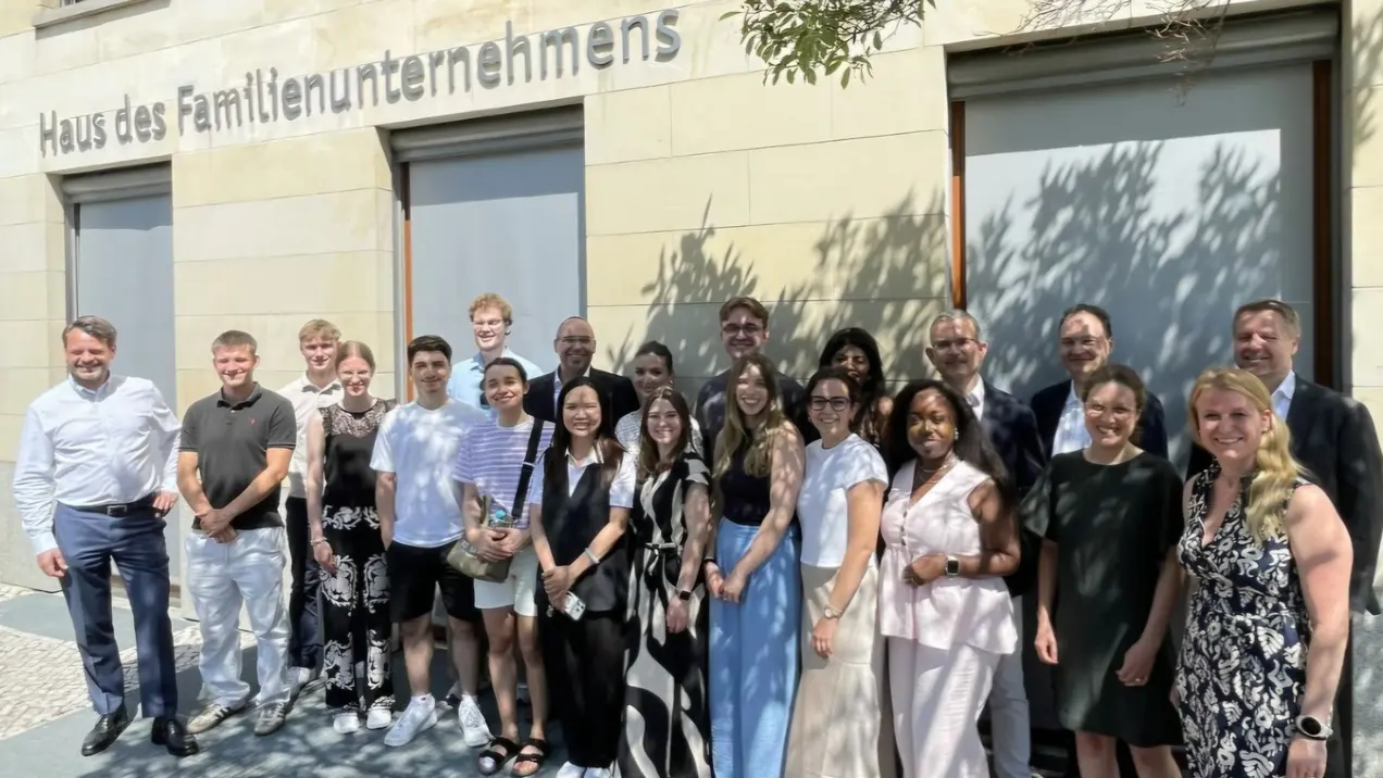 Eine Gruppe von 21 Personen steht vor dem Gebäude "Haus des Familienunternehmens". Die Menschen sind vielfältig gekleidet, lächeln und schauen in die Kamera. Es ist ein sonniger Tag mit Schatten von Bäumen im Vordergrund.