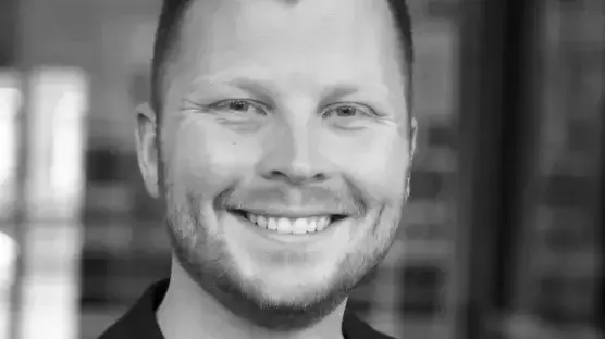 Ein lächelnder Mann mit mittellangen, blonden Haaren und einem gut gepflegten Bart trägt einen schwarzen Blazer und ein T-Shirt. Der Hintergrund ist unscharf und zeigt eine moderne Umgebung.