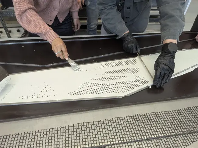 Zwei Personen arbeiten an einer Oberfläche, die mit einer weißen Mischung bestrichen wird. Eine Person hält einen Spachtel, während die andere die Fläche gleichmäßig verteilt. Beide tragen spezielle Handschuhe. Im Hintergrund sind weitere Menschen und Werkzeuge sichtbar.