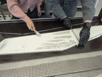 Zwei Personen arbeiten an einer Oberfläche, die mit einer weißen Mischung bestrichen wird. Eine Person hält einen Spachtel, während die andere die Fläche gleichmäßig verteilt. Beide tragen spezielle Handschuhe. Im Hintergrund sind weitere Menschen und Werkzeuge sichtbar.
