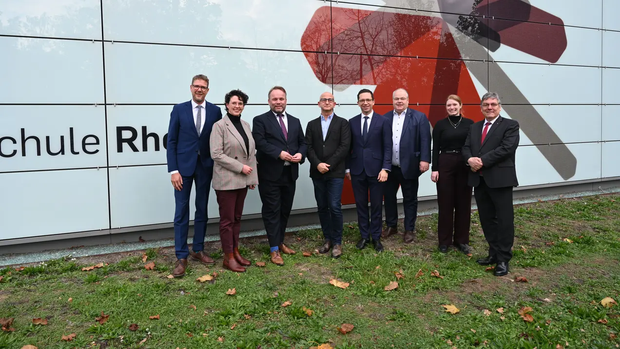 Eine Gruppe von neun Personen steht vor der Fassade einer Schule, die mit einem modernen Design gestaltet ist. Die Menschen sind formell gekleidet und lächeln. Im Hintergrund ist der Schriftzug „Schule Rhein“ zu sehen.