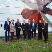 Eine Gruppe von neun Personen steht vor der Fassade einer Schule, die mit einem modernen Design gestaltet ist. Die Menschen sind formell gekleidet und lächeln. Im Hintergrund ist der Schriftzug „Schule Rhein“ zu sehen.