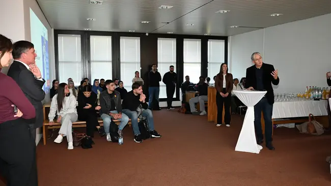 In einem Raum stehen mehrere Personen. Roland Grawinski spricht an einem Stehtisch, während das Publikum gebannt zuhört. Einige Zuschauer sitzen, andere stehen. Der Raum hat große Fenster und eine entspannte Atmosphäre. Im Hintergrund sind Getränke auf einem Tisch sichtbar.