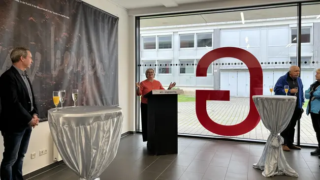 In einem modernen Raum stehen mehrere Personen an Stehtischen. Eine Frau spricht am Podium, während zwei weitere Personen zuhören. Im Hintergrund ist ein großes Fenster mit einer markanten roten Zahl "3" zu sehen. Die Atmosphäre ist festlich und informell.