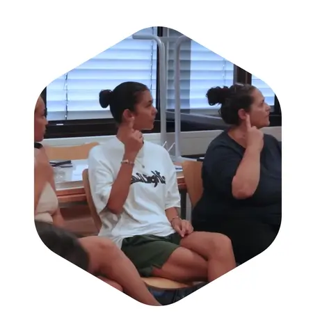 Eine Gruppe von Frauen sitzt in einem Raum. Einige von ihnen zeigen mit den Händen Gesten. Die Frau in der Mitte trägt ein einfarbiges T-Shirt und hat einen Zopf. Der Fokus liegt auf den Gesten und dem Austausch zwischen den Teilnehmerinnen.