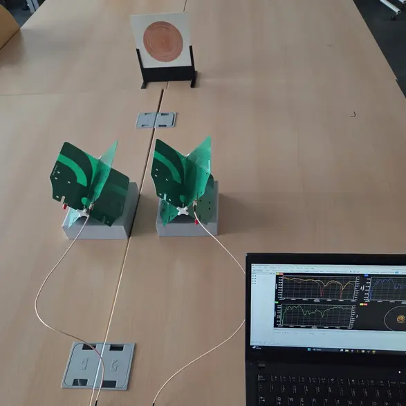 Auf einem Tisch stehen zwei grüne Modelle, die mit Kabeln verbunden sind. Daneben liegt ein Laptop, auf dessen Bildschirm grafische Daten angezeigt werden. Im Hintergrund befindet sich ein Objekt, das in einem Ständer aufgestellt ist.
