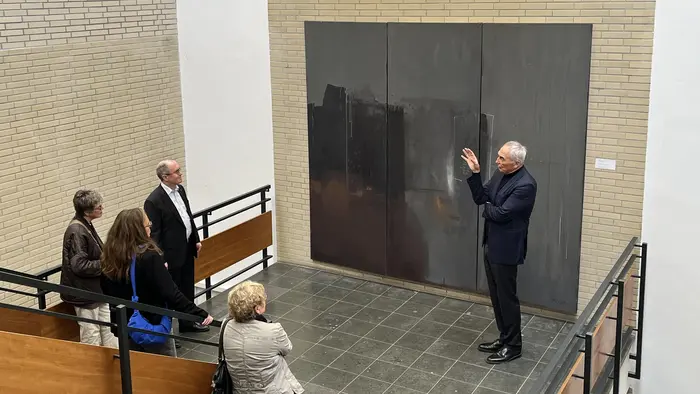 Eine Gruppe von fünf Personen steht in einem Ausstellungsraum und betrachtet ein großes, graues Kunstwerk an der Wand. Ein Mann in einem Anzug erklärt das Kunstwerk gestenreich. Die Atmosphäre ist informativ und kreativ.
