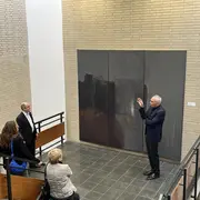 Eine Gruppe von fünf Personen steht in einem Ausstellungsraum und betrachtet ein großes, graues Kunstwerk an der Wand. Ein Mann in einem Anzug erklärt das Kunstwerk gestenreich. Die Atmosphäre ist informativ und kreativ.