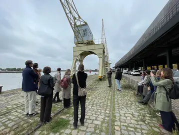 A group of students and their teachers stand on the banks of the River Scheldt in Antwerp, next to a historic crane. The sky is cloudy, and several vehicles and a building are visible in the background. The students are guiding each other through the city and presenting places undergoing change as part of the HERILAND programme.
