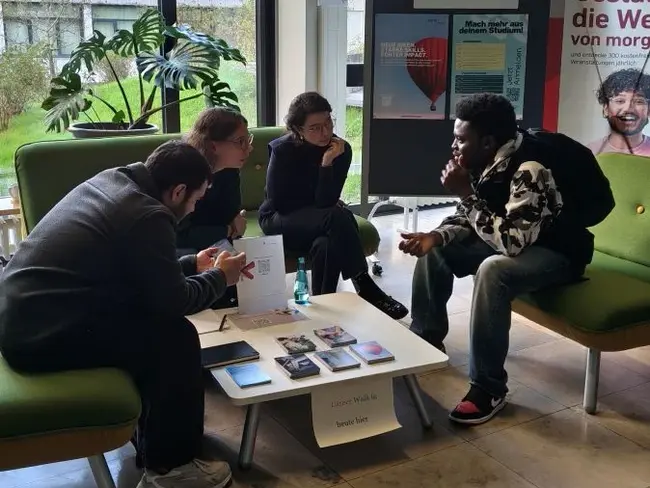 Vier Personen sitzen in einem Aufenthaltsbereich und unterhalten sich. Vor ihnen liegt ein Tisch mit Informationsmaterialien. Im Hintergrund steht ein Plakat mit dem Titel „Career Walk-in“. Die Atmosphäre ist informell und einladend.