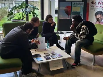 Vier Personen sitzen in einem Aufenthaltsbereich und unterhalten sich. Vor ihnen liegt ein Tisch mit Informationsmaterialien. Im Hintergrund steht ein Plakat mit dem Titel „Career Walk-in“. Die Atmosphäre ist informell und einladend.