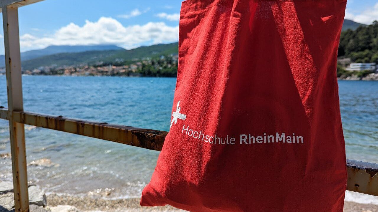An den Strand mit HSRM-Tasche im Auslandssemester in Kroatien, Rijeka Auf dem Bild ist die rote Umhängetasche der HSRM zu sehen, die an einem Geländer hängt. Im Hintergrund ist das Meer zu sehen.