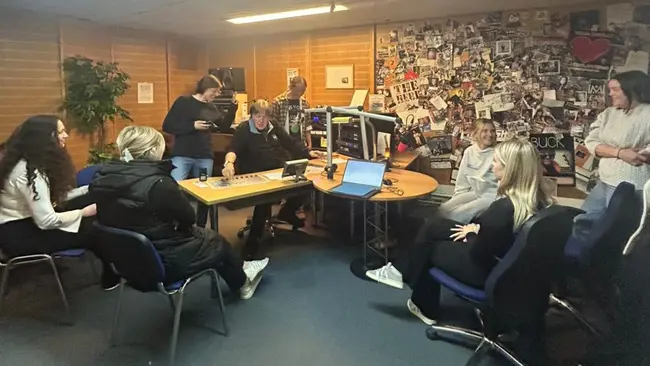 Eine Gruppe von acht Personen sitzt und steht in einem Raum, der mit Plakaten dekoriert ist. Sie arbeiten an Computern und scheinen in ein Gespräch vertieft zu sein. Einige lachen oder schauen auf ihre Handys. Der Raum vermittelt eine kreative und entspannte Atmosphäre.