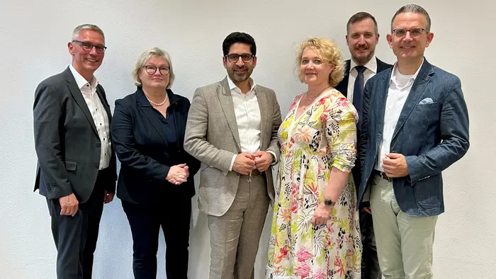 Sechs Personen stehen in einer Reihe vor einer hellen Wand. Sie sind formell gekleidet und lächeln in die Kamera. Die Gruppe umfasst drei Männer und drei Frauen, die miteinander interagieren.