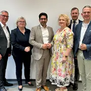 Sechs Personen stehen in einer Reihe vor einer hellen Wand. Sie sind formell gekleidet und lächeln in die Kamera. Die Gruppe umfasst drei Männer und drei Frauen, die miteinander interagieren.