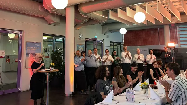 Prof. Dr. Eva Waller steht an einem Tisch und spricht zu einer Gruppe von applaudierenden Personen. Der Raum ist festlich dekoriert, mit Blumen auf den Tischen und runden Lampen an der Decke. 