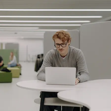 Ein junger Mann mit Brille sitzt an einem Tisch und arbeitet an einem Laptop. Er trägt einen grauen Pullover und konzentriert sich auf den Bildschirm. Im Hintergrund sind einige Menschen zu sehen, die in entspannter Umgebung miteinander sprechen.