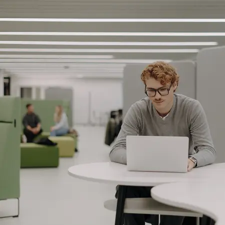 Ein junger Mann mit Brille sitzt an einem Tisch und arbeitet an einem Laptop. Er trägt einen grauen Pullover und konzentriert sich auf den Bildschirm. Im Hintergrund sind einige Menschen zu sehen, die in entspannter Umgebung miteinander sprechen.
