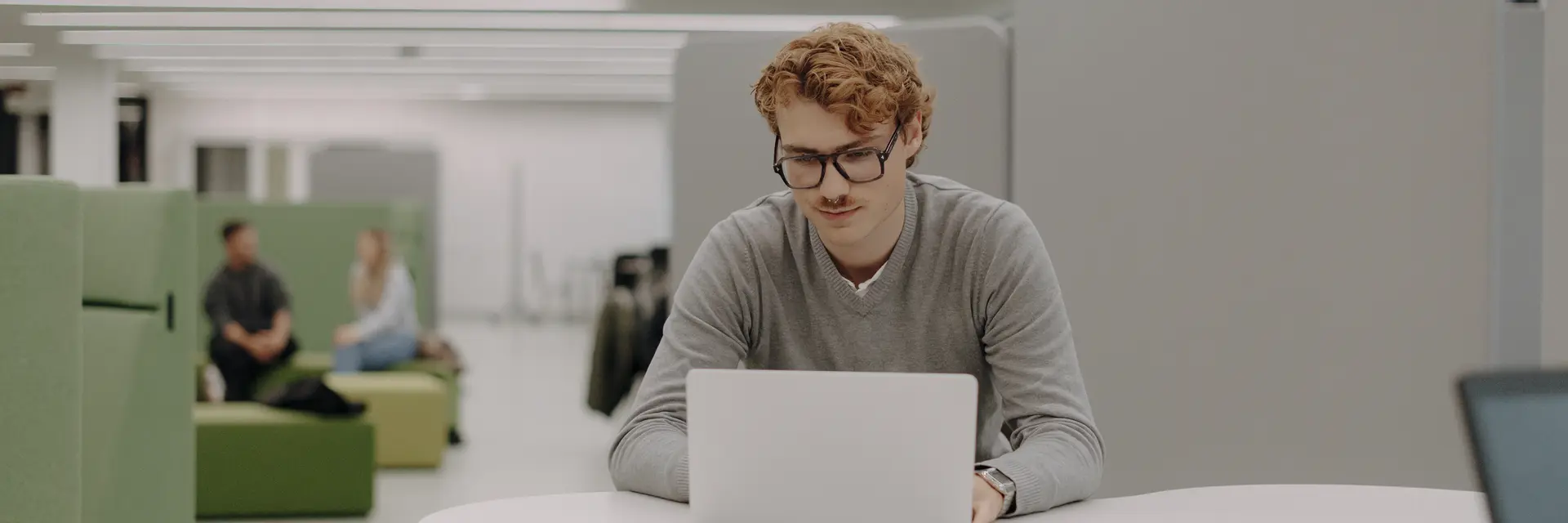 Ein junger Mann mit Brille sitzt an einem Tisch und arbeitet an einem Laptop. Er trägt einen grauen Pullover und konzentriert sich auf den Bildschirm. Im Hintergrund sind einige Menschen zu sehen, die in entspannter Umgebung miteinander sprechen.