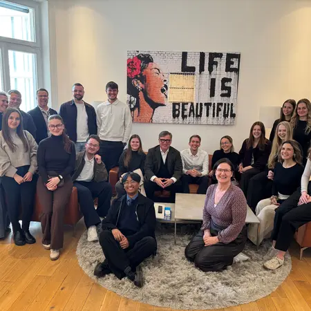 Eine Gruppe von etwa 20 Personen steht und sitzt in einem modernen Raum. Im Hintergrund hängt ein Kunstwerk mit der Aufschrift „LIFE IS BEAUTIFUL“. Die Anwesenden lächeln und sind lässig, in einem freundlichen, professionellen Umfeld.