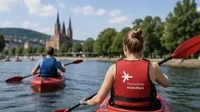 Zwei Personen paddeln in roten Kajaks auf einem ruhigen Fluss. Im Hintergrund sind Bäume und eine Kirche mit hohen Türmen sichtbar. Die Szene vermittelt ein Gefühl von Freizeitaktivität und Natur.