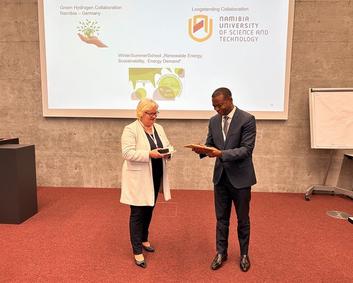 Prof. Dr. Eva Waller, Präsidentin der Hochschule RheinMain, und Dr. Erold Naomab, Vice-Chancellor der Namibia University of Science and Technology, stehen vor einer Präsentationsfolie und tauschen Gastgeschenke aus