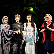 Prof. Dr. Eva Waller, Präsidentin der Hochschule RheinMain, steht mit Schüler:innen der Wilhelm-Heinrich-von-Riehl Schule auf der Bühne des Hessischen Staatstheaters.