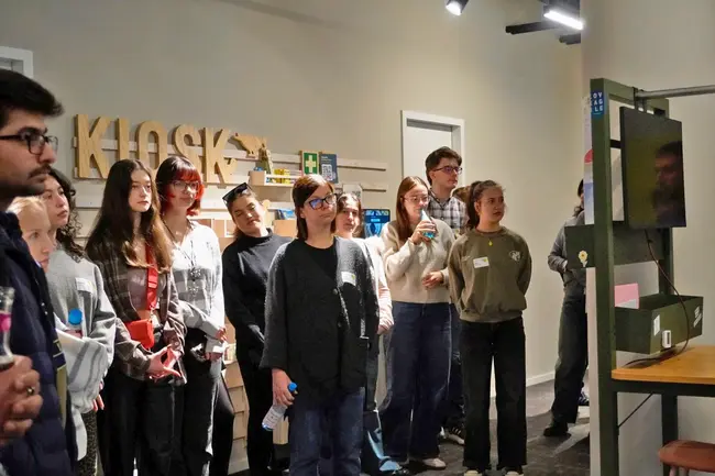 Eine Gruppe von Menschen steht in einem Raum und schaut auf einen Bildschirm. Einige Teilnehmer haben Namensschilder. Im Hintergrund befindet sich eine Wand mit verschiedenen Objekten und dem Schriftzug "KLOS". Die Atmosphäre wirkt aufmerksam und interessiert.