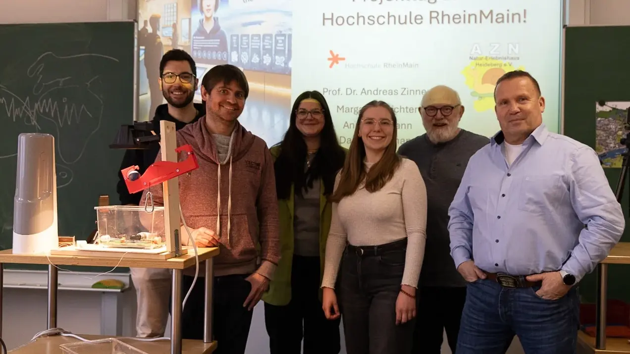 Eine Gruppe von sechs Personen steht lächelnd vor einer Präsentationstafel, die den Schriftzug „Herzlich Willkommen zum Projekttag an der Hochschule RheinMain“ zeigt. Auf einem Tisch sind einige technische Geräte und Materialien zur Demonstration platziert.