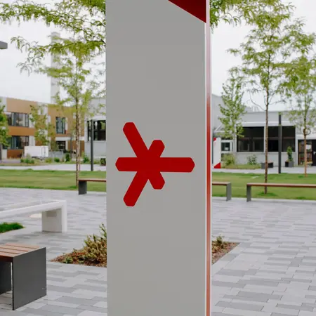 The image shows a modern outdoor area featuring a large white pillar with a red symbol, a bench, and a black seating area. Lush greenery and trees are visible in the background, along with contemporary buildings, creating a welcoming urban space.