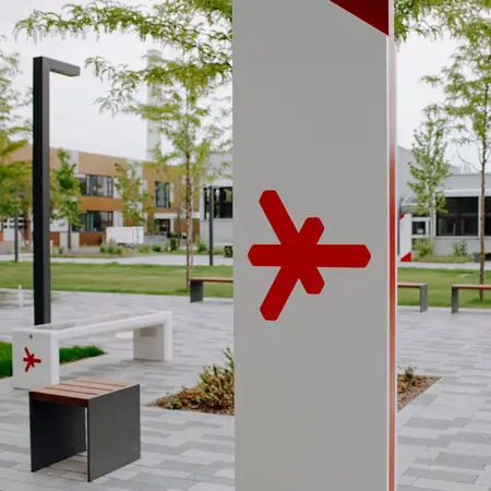 Ein moderner, offener Platz mit Grünflächen und schlichten Sitzgelegenheiten. Im Vordergrund steht ein markanter, weißer Pfeiler mit einem roten Symbol. Umgebende Bäume und Gebäude fügen sich harmonisch in die Landschaft ein, während gepflasterte Wege den Raum strukturieren.