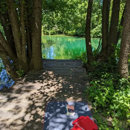 Eine Holzbrücke führt zu einem klaren, grünlichen Gewässer, umgeben von Bäumen. Auf der Brücke liegt eine Yogamatte, neben der ein rotes Handtuch und ein Buch aufgeschlagen sind. Der Schatten der Bäume erzeugt ein ruhiges, einladendes Ambiente.