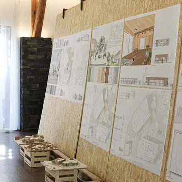 Die Abbildung zeigt eine Wand mit Postern der Pläne und Entwürfe der Studierenden über die Dorfmitte in Wallroth. Unter den Plänen stehen kleine Holzkisten, auf denen sich Architekturmodelle befinden.