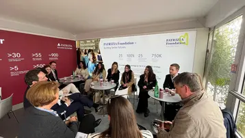 Eine Gruppe von Menschen sitzt an Tischen in einem Raum und diskutiert. Im Hintergrund ist eine Präsentation der Patrizia Foundation zu sehen, die Zahlen und Informationen zur Stiftung enthält.