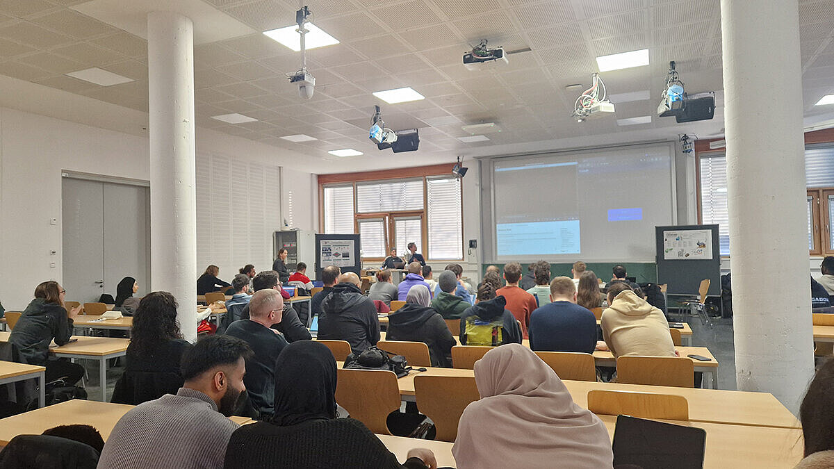 In einem großen Raum sitzen viele Studierende und hören einem Kurzvortrag zu