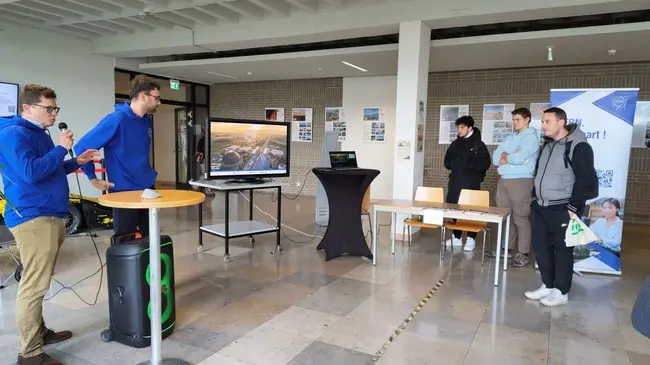Zwei Personen stehen an einem Tisch, einer spricht ins Mikrofon, während der andere an einem Bildschirm mit Landschaftsaufnahmen zeigt. Im Hintergrund sind weitere Personen zu sehen, die der Präsentation zuhören. Es handelt sich um eine Informationsveranstaltung.