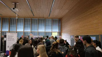 Eine große Gruppe von Menschen versammelt sich in einem Raum, um an einer Veranstaltung teilzunehmen. Im Hintergrund sind Informationsstände zu sehen. Der Fokus liegt auf der Interaktion zwischen den Besuchern und den Informationen, die präsentiert werden.