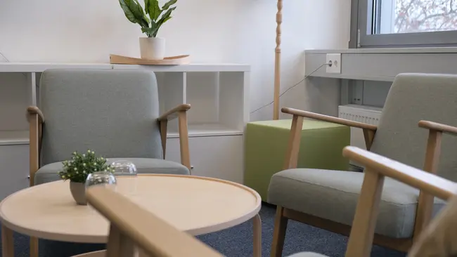 Ein heller Raum mit zwei grauen Sesseln und einem runden Holztisch in der Mitte. Auf dem Tisch befindet sich eine kleine Pflanze und ein Glas. Im Hintergrund steht eine grüne Bank und eine Pflanze auf einem Regal. Das Gesamtbild vermittelt eine einladende Atmosphäre.