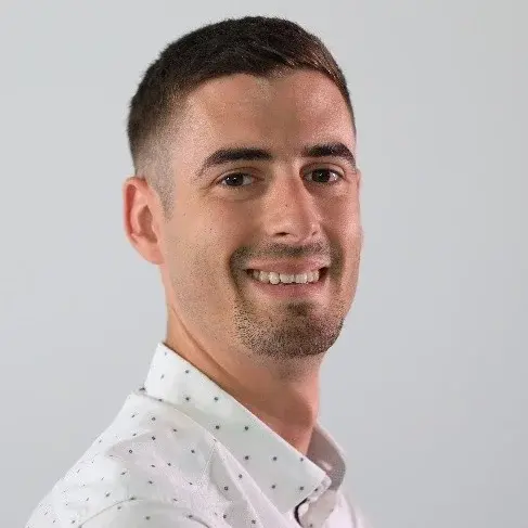 Ein junger Mann mit kurzen, dunklen Haaren und einem freundlichen Lächeln steht vor einem neutralen Hintergrund. Er trägt ein weißes Hemd mit kleinen Punkten und blickt zur Seite.