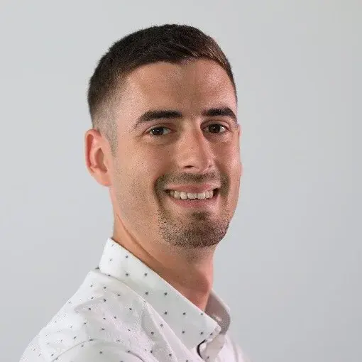 Ein junger Mann mit kurzen, dunklen Haaren und einem freundlichen Lächeln steht vor einem neutralen Hintergrund. Er trägt ein weißes Hemd mit kleinen Punkten und blickt zur Seite.