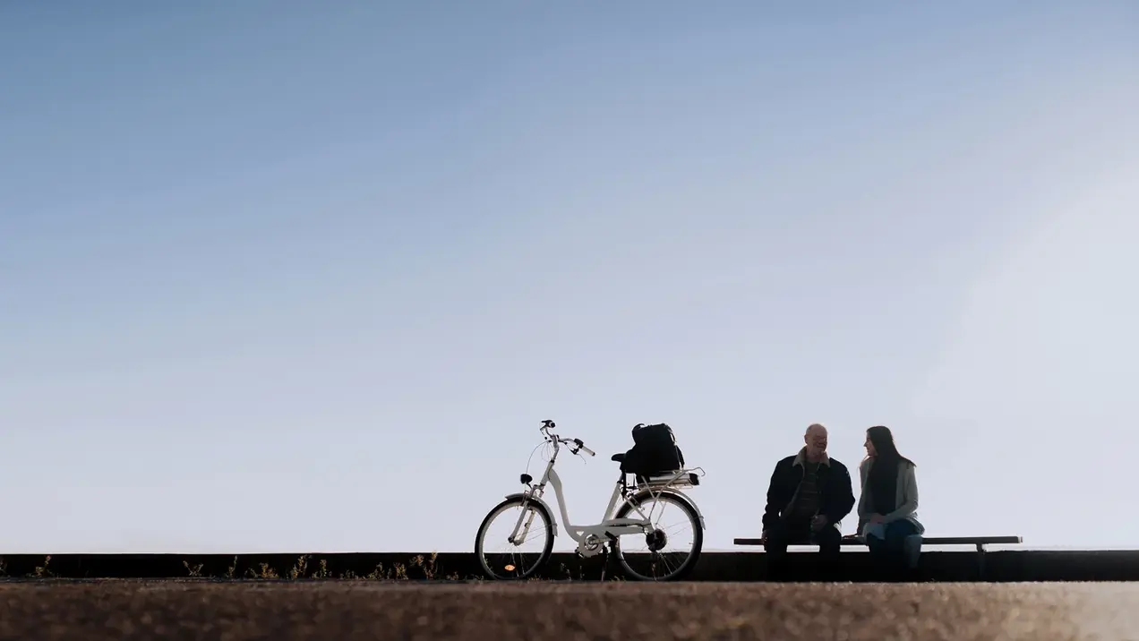 Zwei Personen sitzen auf einer Bank in einer ruhigen Umgebung. Neben ihnen steht ein Fahrrad. Der Himmel ist blau und klar, was eine entspannte Atmosphäre vermittelt.
