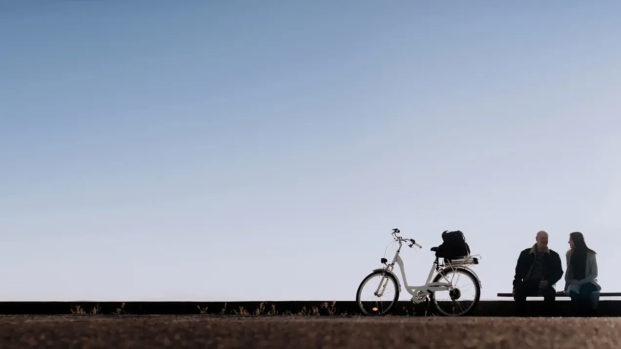 Zwei Personen sitzen auf einer Bank in einer ruhigen Umgebung. Neben ihnen steht ein Fahrrad. Der Himmel ist blau und klar, was eine entspannte Atmosphäre vermittelt.