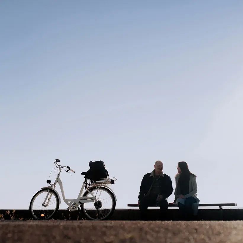 Zwei Personen sitzen auf einer Bank in einer ruhigen Umgebung. Neben ihnen steht ein Fahrrad. Der Himmel ist blau und klar, was eine entspannte Atmosphäre vermittelt.