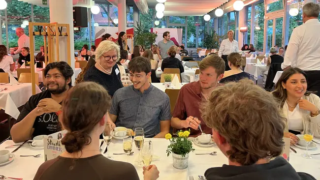 An mehreren Tischen sitzen Studierende in geselliger Runde, während Prof. Dr. Eva Waller Speisen serviert. Die Tische sind mit weißen Tischdecken und Blumen dekoriert, und es gibt Gläser sowie Geschirr.