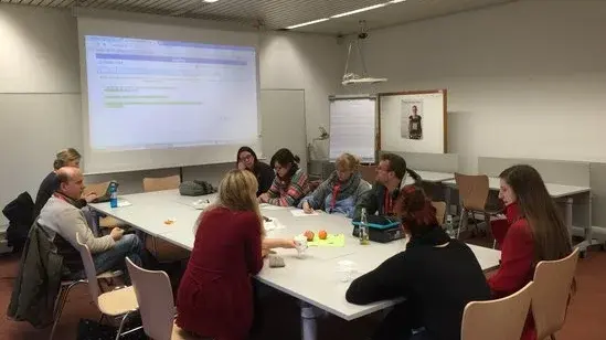 Eine Gruppe, in einer Runde am großen Tisch sitzen, Ideen sammeln und auf eine Leinwand schauen.