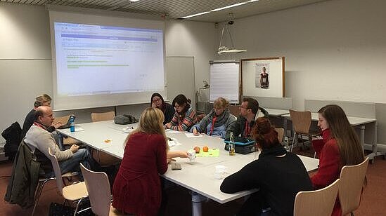 Eine Gruppe, in einer Runde am großen Tisch sitzen, Ideen sammeln und auf eine Leinwand schauen.