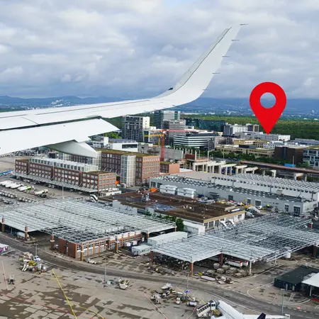 Bildbeschreibung: Eine Flugzeugtragfläche ist im Vordergrund zu sehen, während darunter ein Flughafen und umliegende Gebäude sichtbar sind. Ein rotes Standortsymbol hebt einen bestimmten Punkt auf der Landschaft hervor. Wolken und ein klarer Himmel sind im Hintergrund zu erkennen.